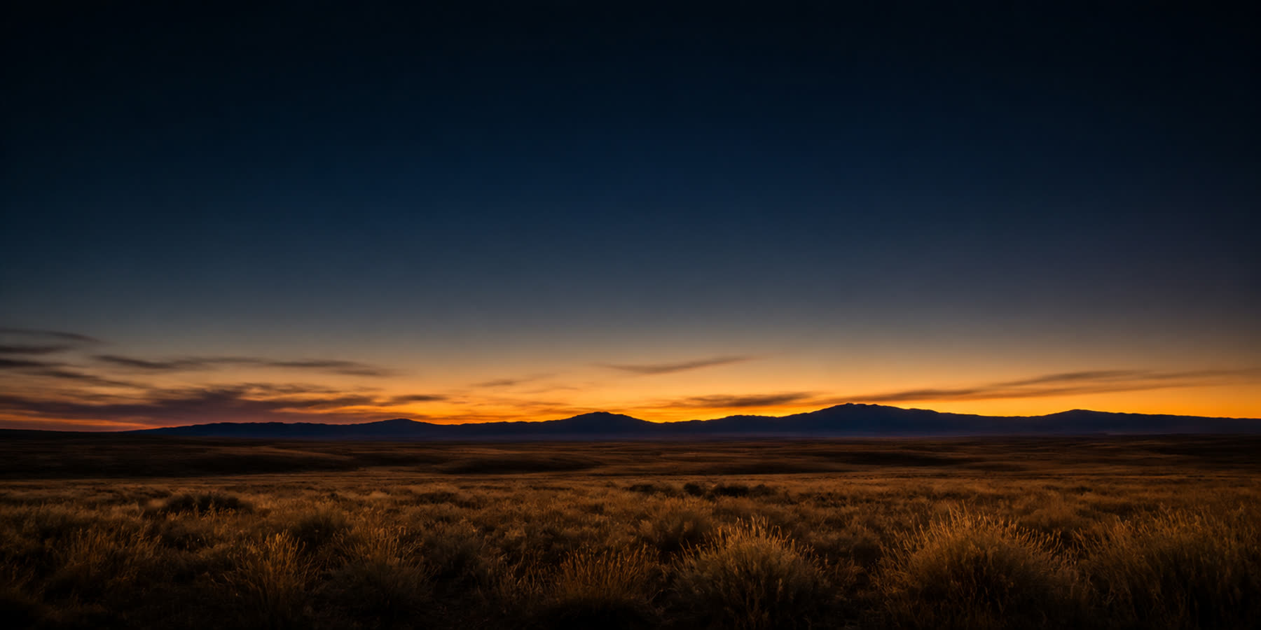 Empty Wyoming high-plains grassland at last light, a dark mountain ridge on the horizon under a narrow amber band and deep blue-black sky, no buildings or people in frame